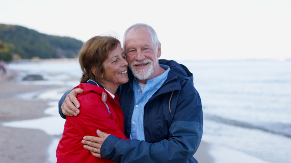 A happy senior couple in love on walk outdoors on beach at morning, hugging and looking at camera.