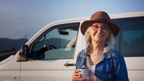 An old woman standing by car, enjoying cup of coffee.