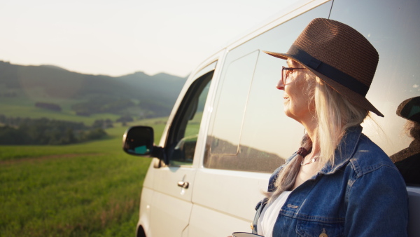 An old woman standing by car, enjoying cup of coffee.