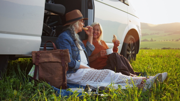 Senior women friends sitting by a car boot outdoors at sunset, caravan trip holiday.