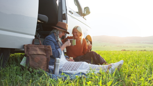 Senior women friends sitting by a car boot outdoors at sunset, caravan trip holiday.