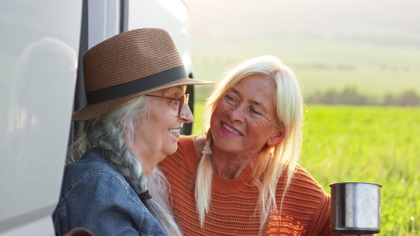 Senior women friends sitting by a car boot outdoors at sunset, caravan trip holiday.