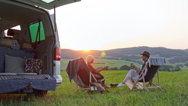 Senior women friends eating and sitting by a car boot outdoors at sunset, caravan trip holiday.