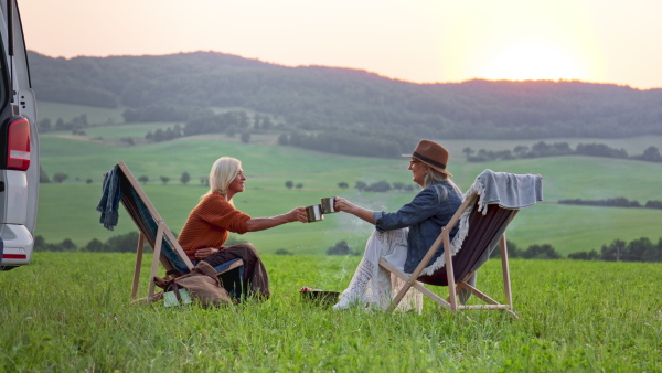 Senior women friends eating and sitting by a car boot outdoors at sunset, caravan trip holiday.