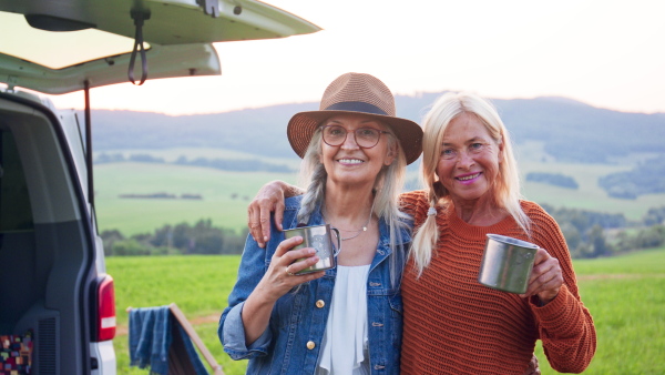 Senior women friends standing by a car outdoors and drinking tea, caravan trip holiday.