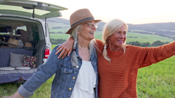 Senior women friends standing by a car outdoors and hugging, caravan trip holiday.