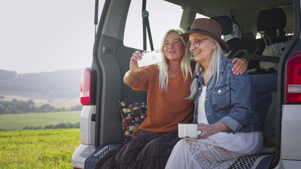 Senior women friends sitting in a car boot and taking selfie outdoors in nature, caravan trip holiday.