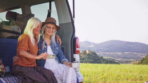 Senior women friends sitting in a car boot and taking selfie outdoors in nature, caravan trip holiday.