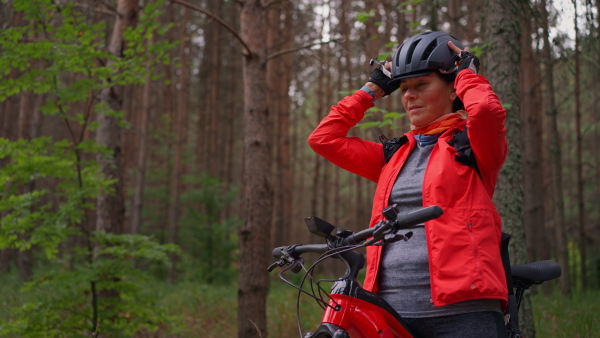 A senior woman biker putting on cycling helmet outdoors in forest in autumn day.