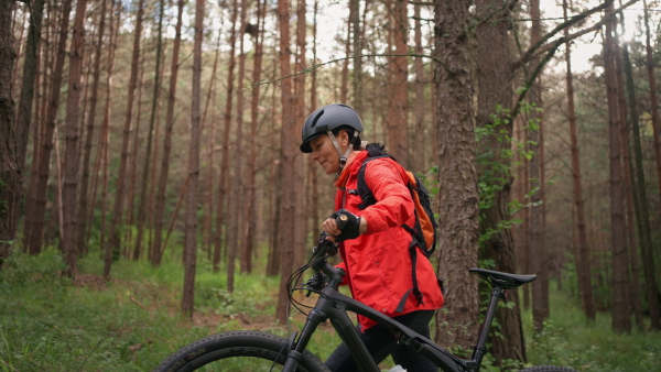 A senior woman biker walking and pushing bike outdoors in forest in autumn day.