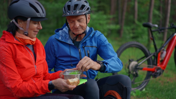 A happy senior couple bikers with eating snack outdoors in forest in autumn day.