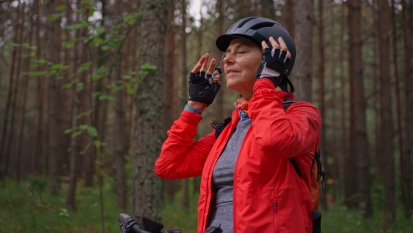 A senior woman biker putting on cycling helmet outdoors in forest in autumn day.