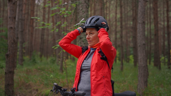 A senior woman biker putting on cycling helmet outdoors in forest in autumn day.