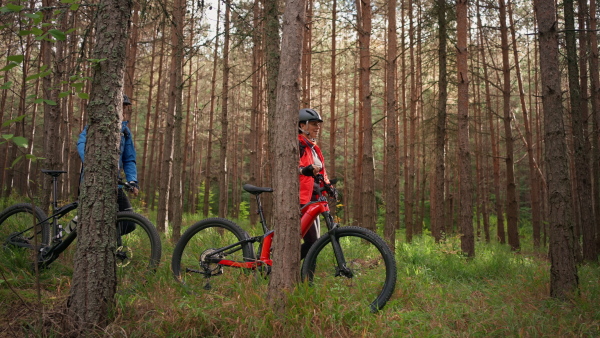 A senior couple bikers walking and pushing e-bikes outdoors in forest in autumn day.