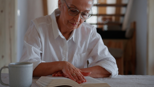 A thoughtful senior woman sitting at table and reading book at home.