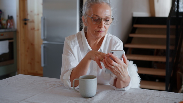 A sad senior woman sitting at table and using smartphone at home.