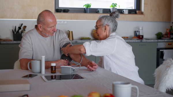 A senior couple spending time together in morning in kitchen, measuring blood pressure.