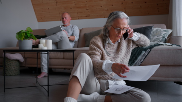 A senior woman holding papers and talking on smartphone near husban using laptop at home