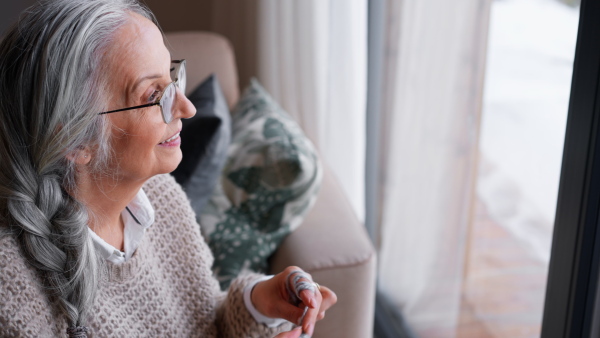 A happy senior woman is looking throug window when knitting at home.