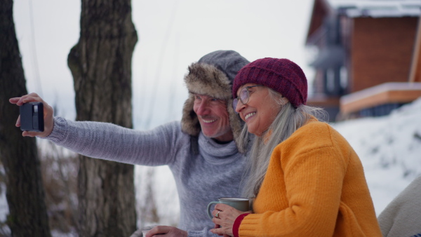 A senior couple taking selfie outdoors on winter holiday in front of cabin.