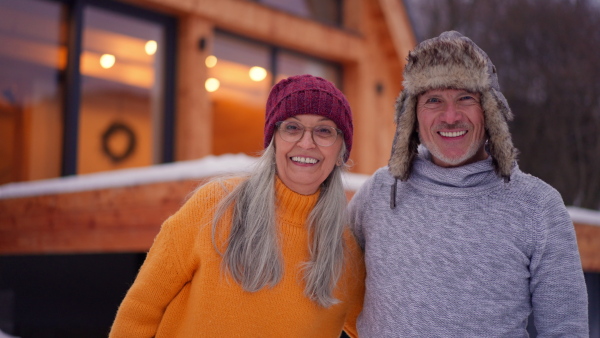 A senior couple looking at camera outdoors on winter holiday in front of cabin.