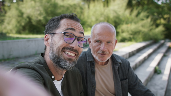 A happy senior man with his mature son sitting on stairs in park and looking at camera.