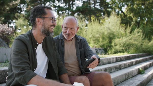 A happy senior man with his mature son sitting on stairs in park and using smartphone.