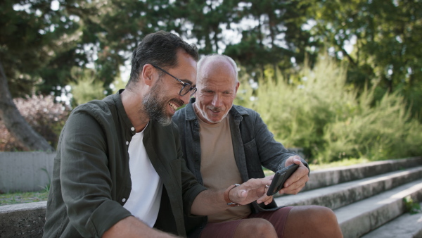 A happy senior man with his mature son sitting on stairs in park and using smartphone.