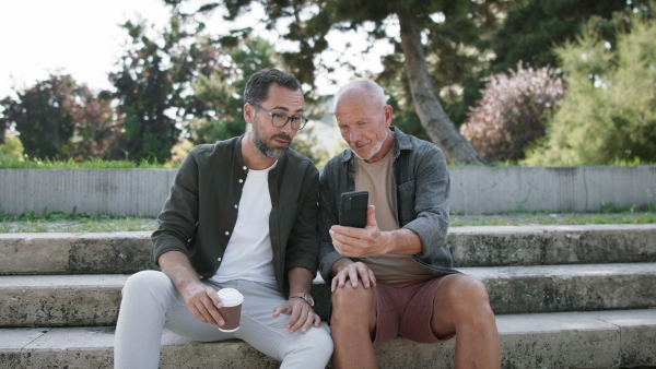 A happy senior man with his mature son sitting on stairs in park and using smartphone.