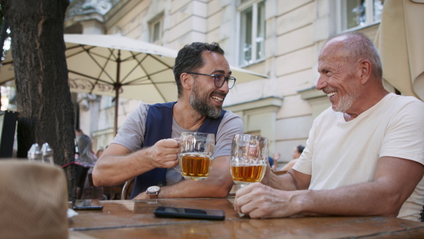 A happy senior man with his mature son drinking beer outdoors in pub.