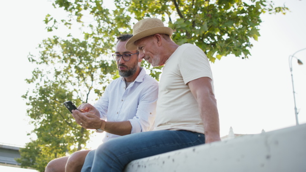 A happy senior man with his mature son sitting on stairs in park and using smartphone.