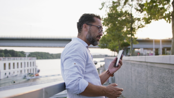 A happy mature man using smartphone and resting outdoors in riverside.