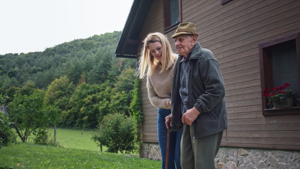 A happy young female caregiver visiting senior man at home, taking patient for walk outdoors in garden.