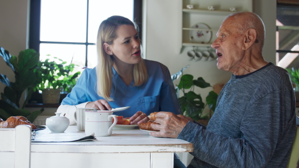 A young female caregiver or healthcare worker visiting senior man indoors at home, assisting with breakfast