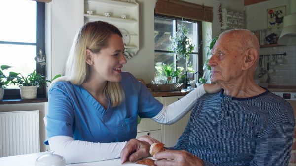 A young female caregiver or healthcare worker visiting senior man indoors at home, assisting with breakfast