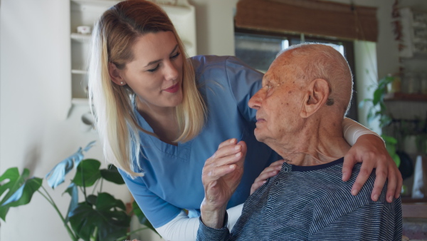 A young female caregiver or healthcare worker visiting senior man indoors at home, talking