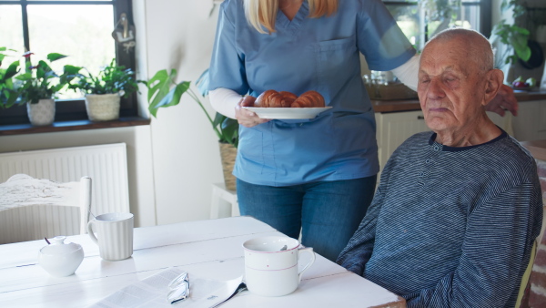 A young female caregiver or healthcare worker visiting senior man indoors at home, assisting with breakfast