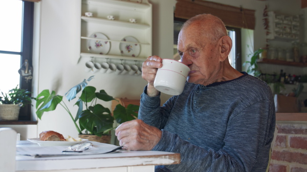 An old senior man sitting, drinking milk and having breakfast indoors at home.