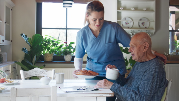 A young female caregiver or healthcare worker visiting senior man indoors at home, assisting with breakfast