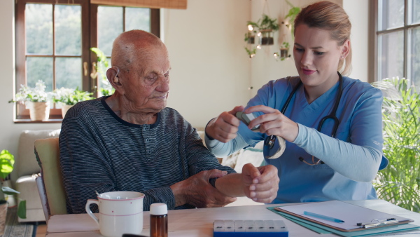 A healthcare worker or caregiver visiting senior man indoors at home, measuring blood pressure.