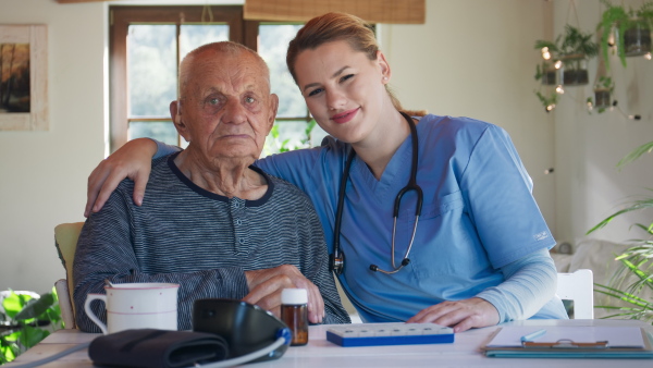 A happy healthcare worker or caregiver visiting senior woman indoors at home, talking and looking at camera.