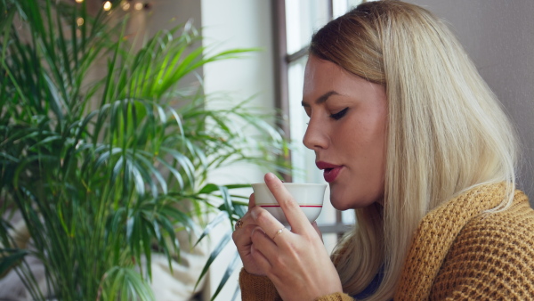 A close up of young woman in sweater holding tea cup and sitting on floor indoors at home.