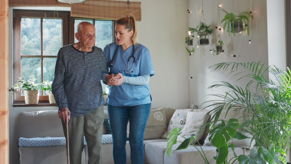 A young female healthcare worker or caregiver visiting senior man indoors at home, holding and walking with patient.