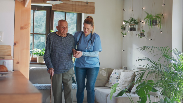 A young female healthcare worker or caregiver visiting senior man indoors at home, holding and walking with patient.