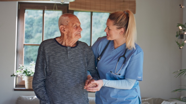 A young female healthcare worker visiting senior man indoors at home, holding and walking with patient.