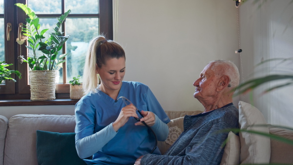 A young female healthcare worker or caregiver visiting senior man indoors at home, examining patient with stethoscope.