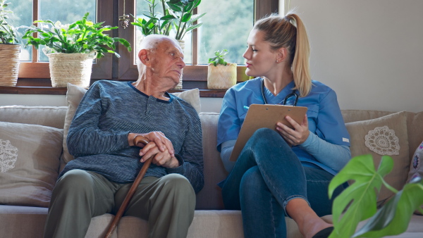 A young female healthcare worker or caregiver visiting senior man indoors at home, examining patient, talking and writing record.