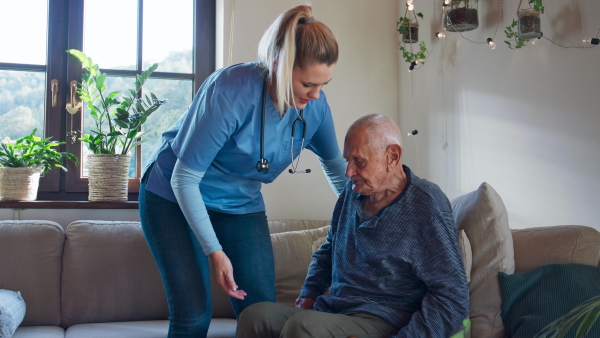 A young female healthcare worker or caregiver visiting senior man indoors at home, helping patient to stand up and walk.