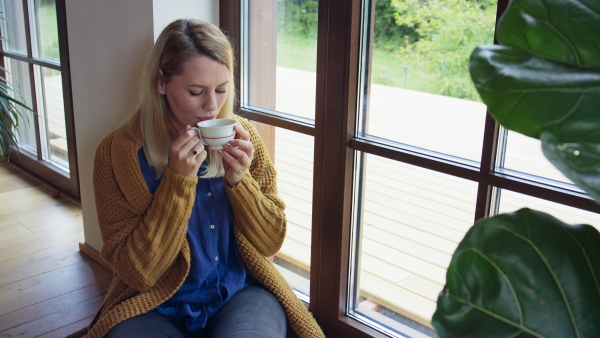 A young woman in sweater drinking tea, sitting on floor and looking out through window indoors at home.