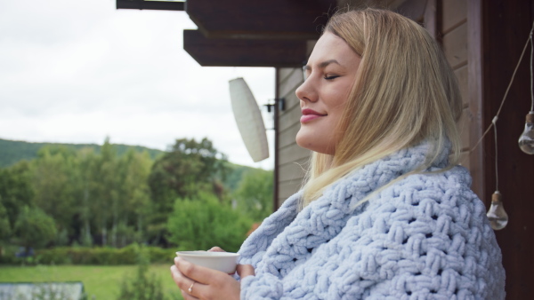 A happy young woman wrapped in blanket drinking tea, relaxing and standing outside on house terrace.